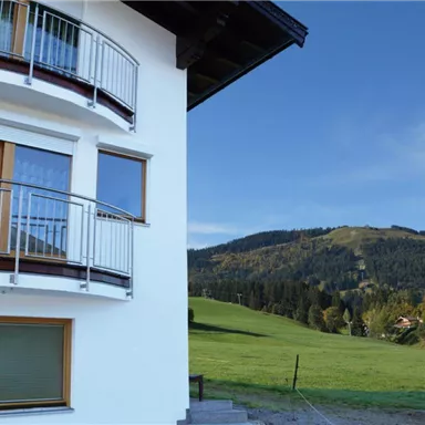 A modern building with a balcony and large windows. In the background, a green meadow landscape extends with gentle hills.