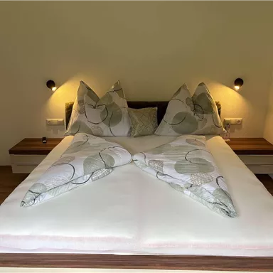 Ein gemütliches Schlafzimmer mit einem großen Bett und dekorativen Kissen. Die Wände sind neutral, und es gibt sanfte Beleuchtung an beiden Seiten des Bettes.