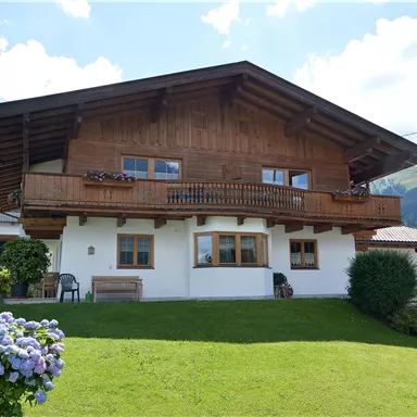 Ein traditionselles alpenländisches Holzhaus mit einem Balkon. Der Garten ist gepflegt und bunte Blumen schmücken den Vordergrund.