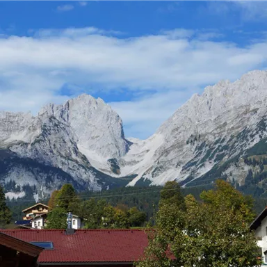 An impressive mountain landscape with tall rocks and a clear blue sky. In the foreground, there are some traditional buildings and trees.
