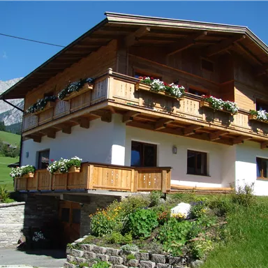 Ein schönes, rustikales Haus mit Holzveranda und Blumenkästen. Die Umgebung ist grün und von Bergen umgeben.