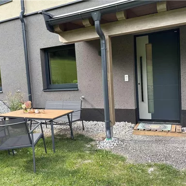 Eine moderne Haustür mit einem kleinen Vorplatz und einem Tisch mit Stühlen daneben. Der Bereich ist von einem gepflegten Garten umgeben.