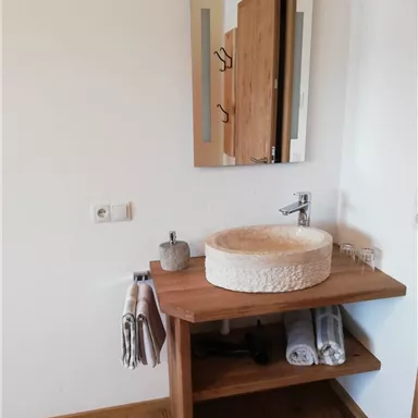 Ein modernes Badezimmer mit einem eleganten Waschbecken aus Stein auf einem Holztisch. Im Hintergrund ist ein Spiegel und Handtücher sind ordentlich aufgehängt.