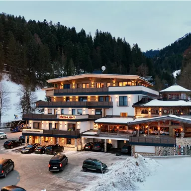 Ein modernes Hotel im Schnee mit umliegenden Bergen. Autos parken vor dem Gebäude, während der Abendhimmel langsam dunkel wird.