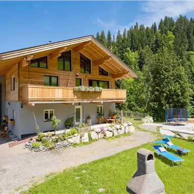 A beautiful wooden house surrounded by green nature. In the foreground, there are lounge chairs and play equipment for children.