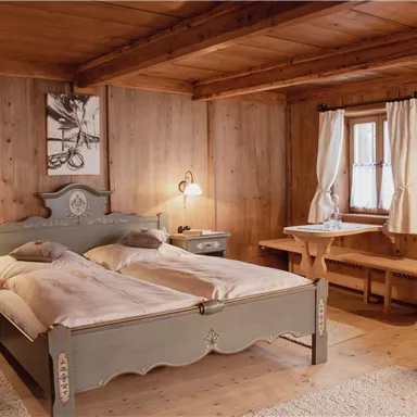 A cozy bedroom with a large, comfortable bed and paneled wooden walls. Next to the bed are a bedside table and a table with chairs.