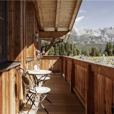 Ein gemütlicher Balkon aus Holz mit einem kleinen Tisch und zwei Stühlen. Im Hintergrund sind beeindruckende Berge und grüne Wälder zu sehen.