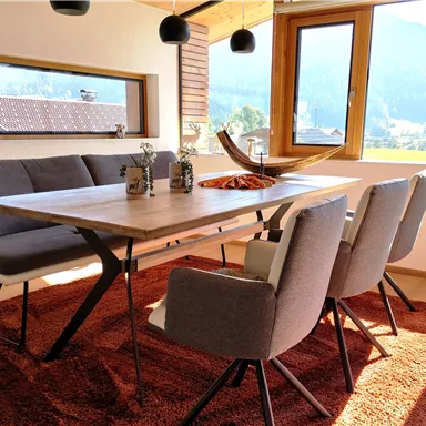 Ein modern eingerichteter Essbereich mit einem großen Tisch und gemütlichen Stühlen. Durch das Fenster sieht man eine malerische Berglandschaft.