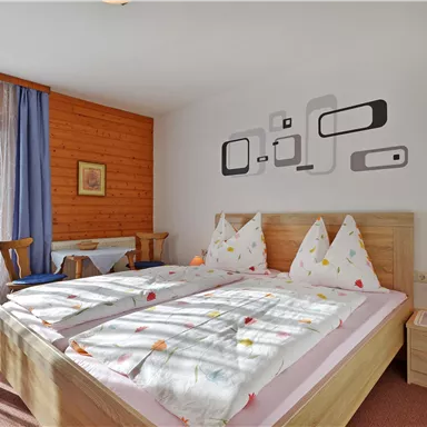 A cozy bedroom with two beds and colorful pillows. The room has large windows and modern wall decor.