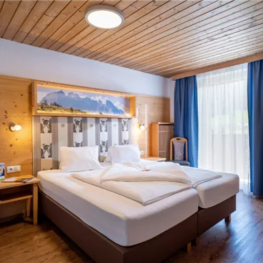 A cozy hotel room with two single beds and wooden decor. There is a large window with blue curtains and a mural of mountains.