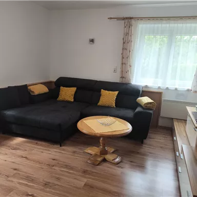 A cozy living room with a black couch and colorful cushions. A round table is in the middle, and a TV is placed on a console.