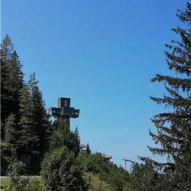 A large cross stands on a hill, surrounded by trees. The clear blue sky creates a calm atmosphere.