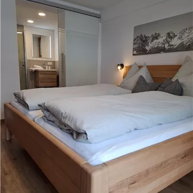 A modern bedroom with a large wooden bed and cozy pillows. In the background, there is a bathroom and a decorative mountain picture.