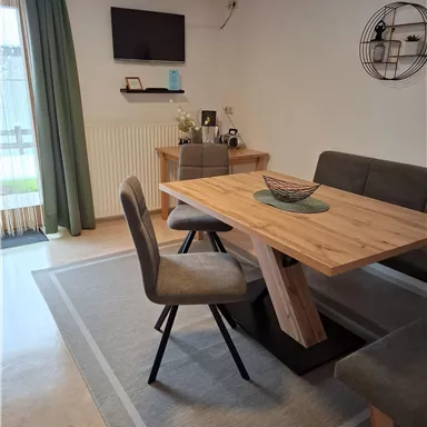 A cozy dining area with a wooden table and modern chairs. In the background, there is a TV and a decorative wall shelf.