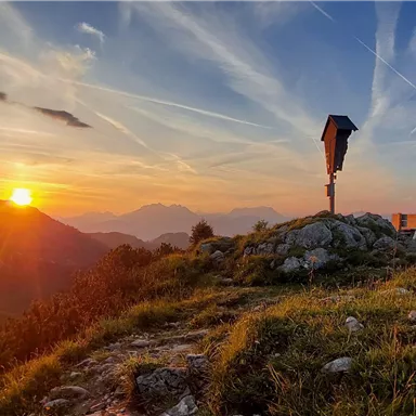 Ein wunderschöner Sonnenuntergang über den Bergen mit bunten Wolken am Himmel. Im Vordergrund sind grüne Wiesen und Felsen zu sehen.