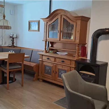 Ein gemütlicher Speisebereich mit einem Holztisch und Stühlen. Im Hintergrund steht ein traditionelles Geschirrschrank und ein Kaminofen.