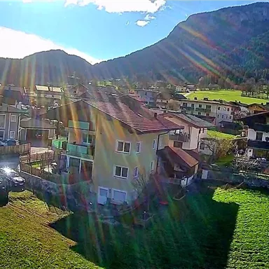 Eine malerische Landschaft mit Häusern und grünen Wiesen, umgeben von Bergen. Die Sonne scheint und sorgt für einen strahlenden Himmel.