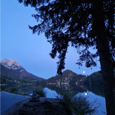A tranquil landscape with a lake, surrounded by mountains and tall trees. The moon reflects gently on the water under a clear, blue sky.