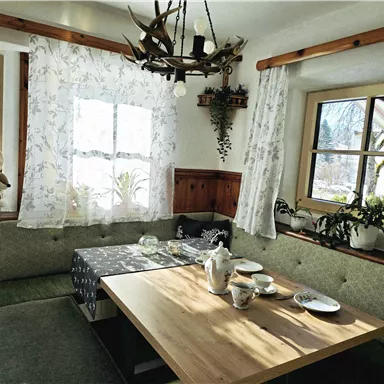 A cozy dining area with wood paneling and windows with curtains. The table is set and surrounded by seating, while plants create a harmonious atmosphere.