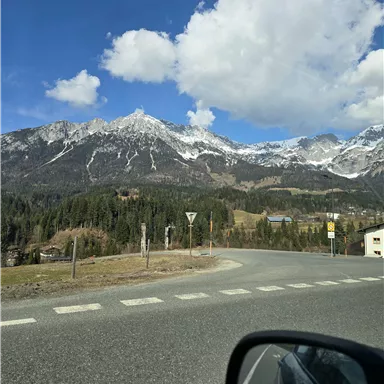 A winding road leads to snow-covered mountains under a clear blue sky. In the foreground, a car mirror is visible.
