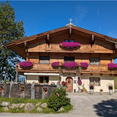 Ein traditionelles Holzhaus mit blühenden Balkonkästen in lila. Es steht in einer malerischen Landschaft unter einem blauen Himmel.
