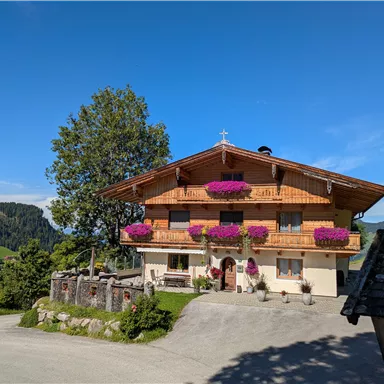Ein schönes, traditionelles altes Haus mit Balkonen voller bunter Blumen. Die umliegende Landschaft ist hügelig und grün unter einem klaren blauen Himmel.
