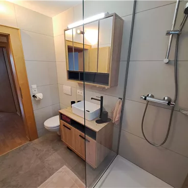 Ein modernes Badezimmer mit einer gläsernen Dusche und einem schlichten Waschtisch. Die Wände sind hell gefliest, und es gibt Holzdetails für einen warmen Akzent.