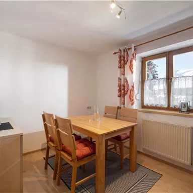 Ein helles Esszimmer mit einem Holzstuhl und einem Tisch. Das Fenster lässt viel Licht herein und ist mit bunten Vorhängen dekoriert.