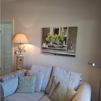 Ein gemütliches Wohnzimmer mit einem hellen Sofa und dekorativen Kissen. An der Wand hängt ein Bild mit einer Fensterbank und Pflanzen.