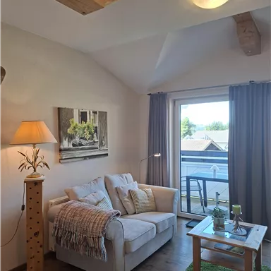 Ein gemütliches Wohnzimmer mit einem beigen Sofa und einer Stehlampe. Große Fenster bieten einen Blick nach draußen und bringen viel Licht in den Raum.