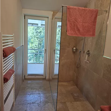 Ein modernes Badezimmer mit einer Glasdusche und zwei Fenstern. Handtücher in sanften Rosatönen sind ordentlich auf einem Handtuchhalter angeordnet.