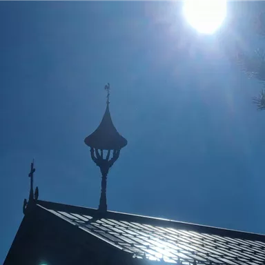 A sunny sky above a small, traditional building with a characteristic roof. A weather vane on the tower and some branches in the foreground are visible.