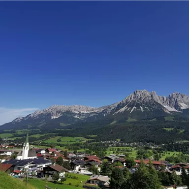 A picturesque Alpine landscape with a small community in the foreground and majestic mountains in the background. The clear blue sky creates a peaceful atmosphere.