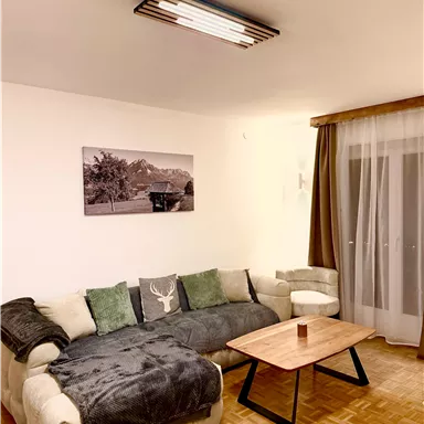 A cozy living room with a large sofa, a coffee table, and a picture on the wall. The windows are decorated with curtains, and the room has warm lighting.
