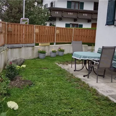 Ein gepflegter Garten mit einer grünen Rasenfläche und bunten Blumen. Es gibt einen Tisch mit Stühlen und einen Sonnenschirm in der Nähe einer Mauer.