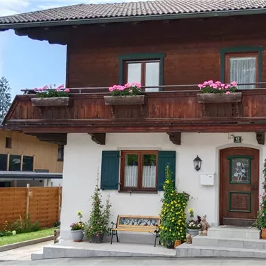Ein traditionelles Haus mit einem Holz-Balkon, der mit Blumen geschmückt ist. Im Vordergrund stehen Pflanzen und eine Bank.