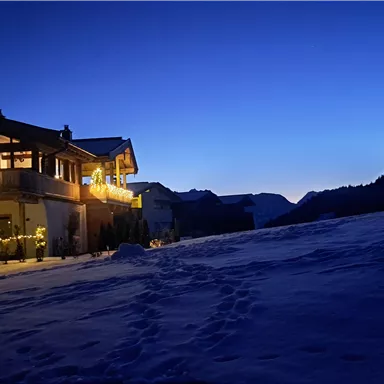 A cozy wooden house in the snow, illuminated by warm light. The sky is clear and shows a beautiful, deep blue shade.