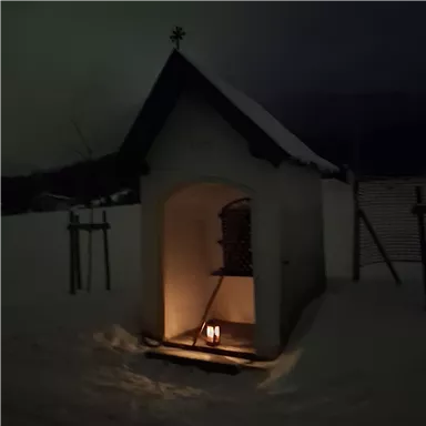 A small chapel in the darkness, surrounded by snow. Inside, a lantern glows, creating a cozy atmosphere.