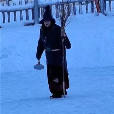 A person in dark clothing is standing on an ice-covered surface. They are holding a broom and a round object in their hand.