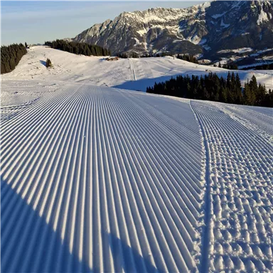 A snowy landscape with well-groomed slopes and majestic mountains in the background. The sky is clear and the sun is shining on the snow.