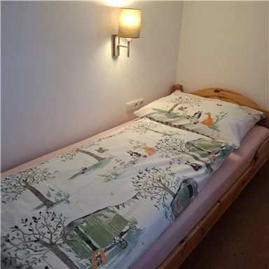 A simple single bed with a colorful duvet and pillow. The wall is painted light, and there's a small lamp above the bed.