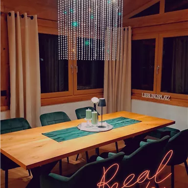 A modern dining room with a long wooden table and stylish green chairs. Above the table hangs a striking pendant lamp with chains.