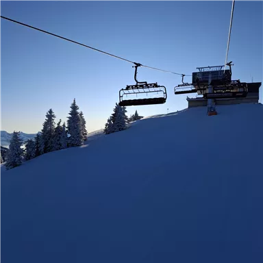 A snow-covered landscape with a ski lift leading into the blue sky. In the background, wintery trees can be seen.