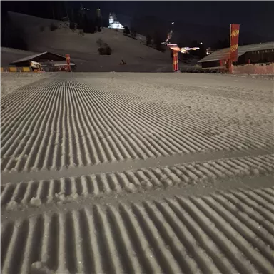 A snowy area with nearly perfect snowcat tracks. In the background, huts and lights are visible, creating a cozy atmosphere.