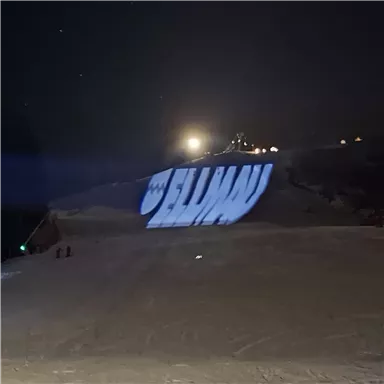 A bright projection of the word "Ellen" on a snowy slope at night. In the background, lights and a snow-covered hill are visible.