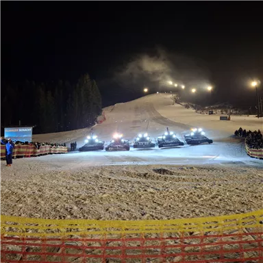 A lighted ski slope at night with several piste grooming vehicles. In the background, spectators can be seen watching the event.