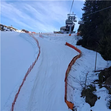 A snowy ski slope with a ski lift in the background. The sky is clear and blue, and there are safety barriers in front of the slope.
