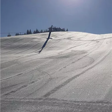 A snowy mountain with ski tracks and radiant sunshine. The sky is clear and blue.