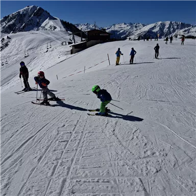 A snow-covered landscape with skiing individuals. In the background, mountains and a blue sky are visible.