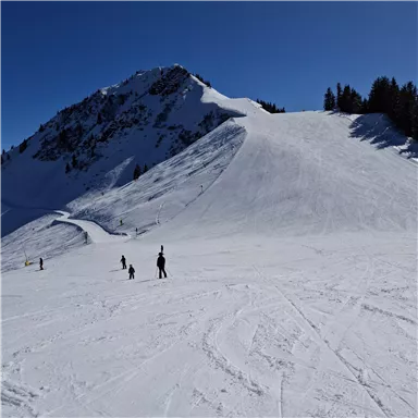 A snowy mountain landscape with skiers in the foreground. The sky is clear and blue.